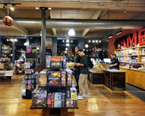 The image depicts the interior of a board game store with shelves filled with various games. Two customers stand near the counter, examining a game, while a cashier is present at the register. The warm lighting and wooden floors give the store a cozy atmosphere.