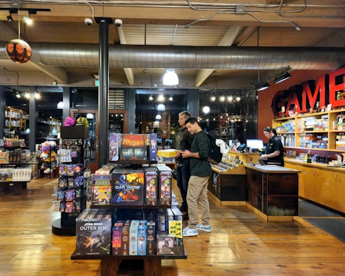 The image depicts the interior of a board game store with shelves filled with various games. Two customers stand near the counter, examining a game, while a cashier is present at the register. The warm lighting and wooden floors give the store a cozy atmosphere.