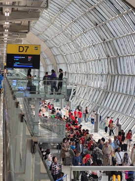 A spacious airport terminal with a high arched ceiling made of glass panels. The terminal is bustling with people, some sitting and others walking with luggage. A digital sign displaying gate information marked D7 is visible. Most people appear relaxed, some engaged in their phones, with a prominent group in red shirts.