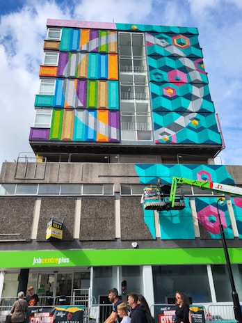 A vibrant mural covers the upper section of a multi-story building. The artwork features geometric patterns and a variety of bright colors including teal, pink, yellow, and orange. Below, a green sign indicates a job center. People are visible at ground level, walking past the building. A person is elevated on a cherry picker, possibly engaged in work on the mural.