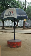 A large outdoor umbrella with a logo and text, positioned in a park setting. The umbrella has a blue canopy with white text and is supported by a central pole anchored in a red circular base. Trees and a paved walkway are visible in the background.