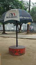 A large outdoor umbrella with a logo and text, positioned in a park setting. The umbrella has a blue canopy with white text and is supported by a central pole anchored in a red circular base. Trees and a paved walkway are visible in the background.