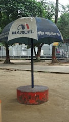A large outdoor umbrella with a logo and text, positioned in a park setting. The umbrella has a blue canopy with white text and is supported by a central pole anchored in a red circular base. Trees and a paved walkway are visible in the background.