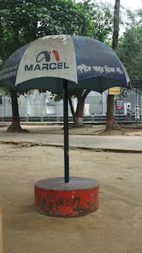 A large outdoor umbrella with a logo and text, positioned in a park setting. The umbrella has a blue canopy with white text and is supported by a central pole anchored in a red circular base. Trees and a paved walkway are visible in the background.