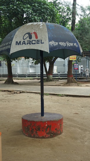 A large outdoor umbrella with a logo and text, positioned in a park setting. The umbrella has a blue canopy with white text and is supported by a central pole anchored in a red circular base. Trees and a paved walkway are visible in the background.