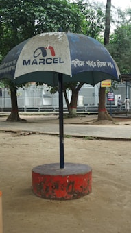 A large outdoor umbrella with a logo and text, positioned in a park setting. The umbrella has a blue canopy with white text and is supported by a central pole anchored in a red circular base. Trees and a paved walkway are visible in the background.