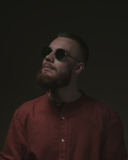A person with a beard wears sunglasses and a dark red shirt, gazing upward against a dark background, creating a mysterious atmosphere.