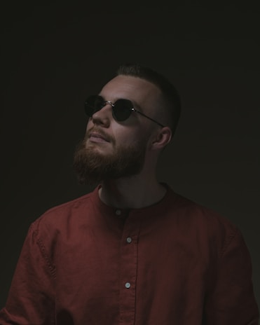 A person with a beard wears sunglasses and a dark red shirt, gazing upward against a dark background, creating a mysterious atmosphere.
