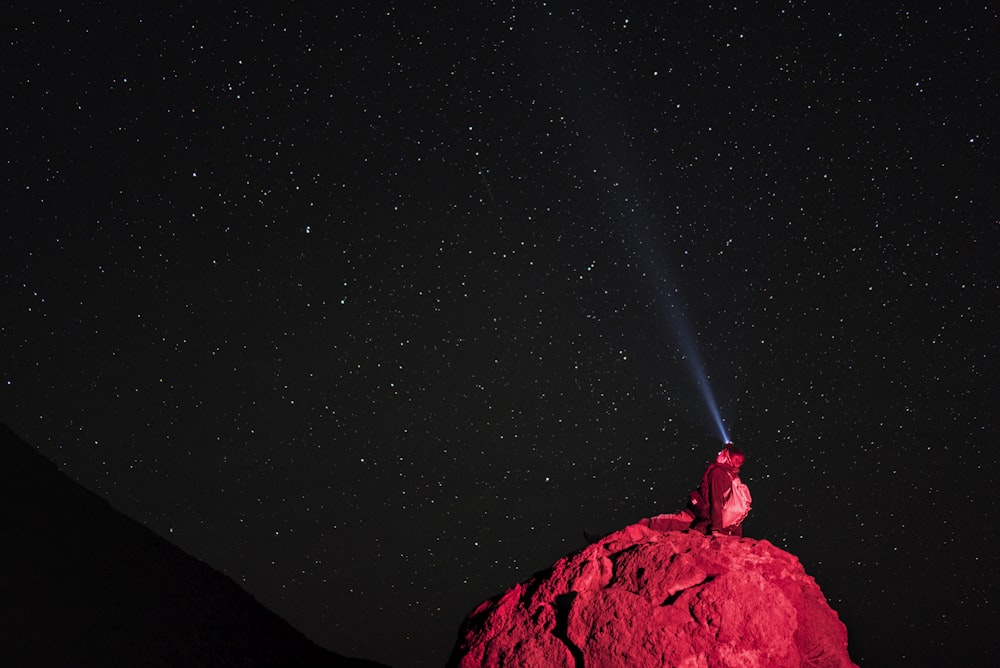 uma pessoa em pé no topo de uma grande rocha sob um céu cheio de estrelas