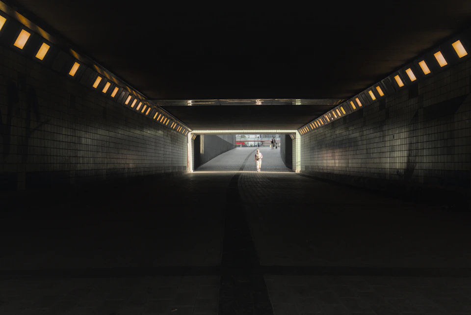 Person walking through underground tunnel illuminated by bright lights casting long shadows everywhere