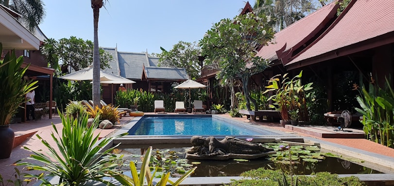 A serene courtyard featuring a pool surrounded by tropical plants and traditional wooden buildings with sloping roofs. Sun loungers and umbrellas line one side of the pool, while a small pond with lotus plants and a statue is in the foreground. The setting is lush with green foliage and ample sunlight.