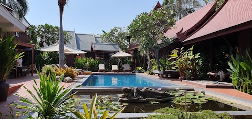 A serene courtyard featuring a pool surrounded by tropical plants and traditional wooden buildings with sloping roofs. Sun loungers and umbrellas line one side of the pool, while a small pond with lotus plants and a statue is in the foreground. The setting is lush with green foliage and ample sunlight.