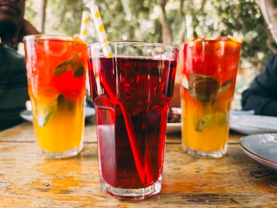 Three glasses of colorful beverages are arranged on a rustic wooden table. The front glass contains a deep red drink, while the two glasses behind it contain vibrant orange-yellow drinks with green mint leaves and slices of fruit visible. The scene is set outdoors with blurred green foliage in the background, evoking a relaxed, refreshing atmosphere.