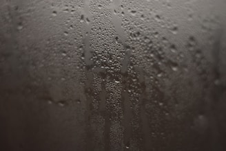 Condensation droplets form on a fogged glass surface, creating a pattern of streaks and water beads. The droplets vary in size and give the surface a textured appearance.