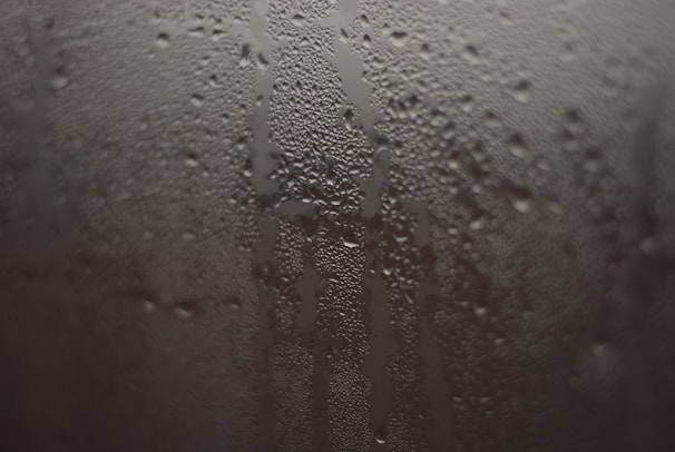 Condensation droplets form on a fogged glass surface, creating a pattern of streaks and water beads. The droplets vary in size and give the surface a textured appearance.