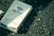 A pack of cigarettes labeled 'Hope' lies on a rough, textured asphalt surface. The packaging includes Japanese text and a graphic design featuring a blue triangle. Lighting is natural with a slightly greenish tint, creating a muted and gritty atmosphere.