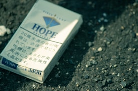 A pack of cigarettes labeled 'Hope' lies on a rough, textured asphalt surface. The packaging includes Japanese text and a graphic design featuring a blue triangle. Lighting is natural with a slightly greenish tint, creating a muted and gritty atmosphere.