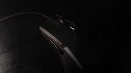 A black gaming mouse with an angular design rests on a dark surface. The mouse features several buttons and a textured grip. The lighting is low, creating a dramatic and mysterious atmosphere.