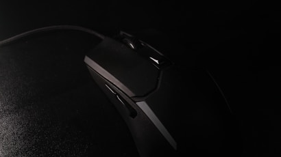 A black gaming mouse with an angular design rests on a dark surface. The mouse features several buttons and a textured grip. The lighting is low, creating a dramatic and mysterious atmosphere.
