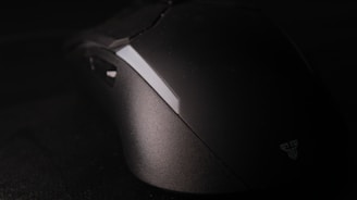 A modern, sleek computer mouse sits on a dark surface. The mouse has a matte finish with a subtle logo on the side. The lighting highlights the contours and enhances the elegant design.