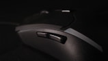 A close-up view of a black gaming mouse with multiple buttons and a textured surface, set against a dark background. The focus is on the side buttons, highlighting the ergonomic design of the device.