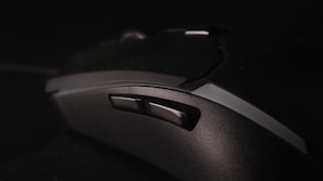 A close-up view of a black gaming mouse with multiple buttons and a textured surface, set against a dark background. The focus is on the side buttons, highlighting the ergonomic design of the device.