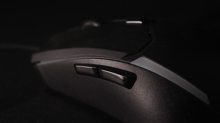 A close-up view of a black gaming mouse with multiple buttons and a textured surface, set against a dark background. The focus is on the side buttons, highlighting the ergonomic design of the device.