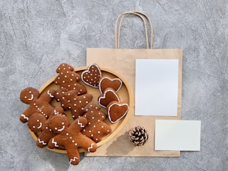 A collection of gingerbread cookies, shaped like gingerbread people and hearts, is arranged on a wooden plate. Beside the plate is a brown paper bag with handles, and two blank white sheets of paper. A pinecone lies near the bottom of the setup, all placed against a textured gray surface.