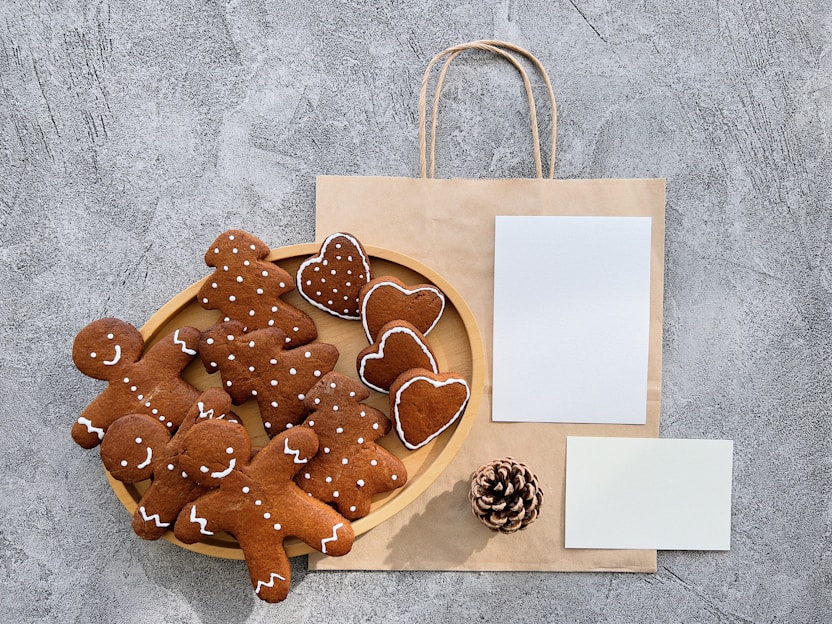 A collection of gingerbread cookies, shaped like gingerbread people and hearts, is arranged on a wooden plate. Beside the plate is a brown paper bag with handles, and two blank white sheets of paper. A pinecone lies near the bottom of the setup, all placed against a textured gray surface.