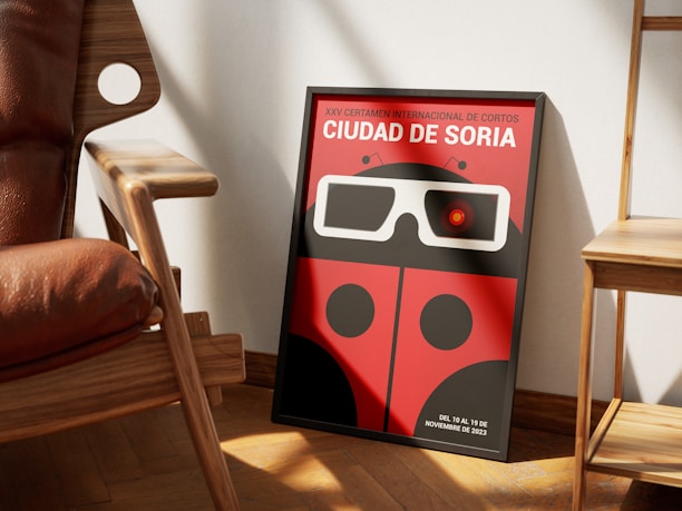 A framed poster leaning against a wall, featuring a stylized ladybug with 3D glasses. The background of the poster is red, and it promotes an international film event, 'Ciudad de Soria,' scheduled for November 2023. The scene includes a wooden chair and a small wooden table, both casting shadows on the wooden floor.