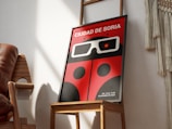 A framed poster featuring a red and black design reminiscent of a ladybug with large stylized 3D glasses. The text at the top is in Spanish, indicating an international short film festival named 'Ciudad de Soria'. The poster is positioned on a wooden chair in a well-lit room with shadows falling across the wall. Beside the chair, there is a portion of a woven wall hanging visible.