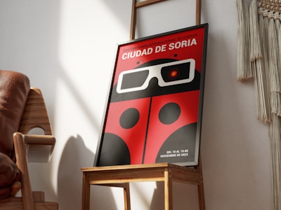 A framed poster featuring a red and black design reminiscent of a ladybug with large stylized 3D glasses. The text at the top is in Spanish, indicating an international short film festival named 'Ciudad de Soria'. The poster is positioned on a wooden chair in a well-lit room with shadows falling across the wall. Beside the chair, there is a portion of a woven wall hanging visible.