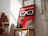 A framed poster featuring a red and black design reminiscent of a ladybug with large stylized 3D glasses. The text at the top is in Spanish, indicating an international short film festival named 'Ciudad de Soria'. The poster is positioned on a wooden chair in a well-lit room with shadows falling across the wall. Beside the chair, there is a portion of a woven wall hanging visible.