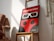 A framed poster featuring a red and black design reminiscent of a ladybug with large stylized 3D glasses. The text at the top is in Spanish, indicating an international short film festival named 'Ciudad de Soria'. The poster is positioned on a wooden chair in a well-lit room with shadows falling across the wall. Beside the chair, there is a portion of a woven wall hanging visible.