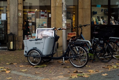 A cargo bike with a large front compartment is parked on a cobblestone street near a storefront with books and other items displayed. There are additional bicycles parked close by, and fallen leaves are scattered on the ground.
