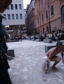 A man with knee and elbow bands is crouching on a paved urban square surrounded by modern and older brick buildings. A crowd of onlookers is gathered around the area, and another person stands in the background wearing red pants, suggesting a performance or public event.