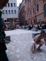 A man with knee and elbow bands is crouching on a paved urban square surrounded by modern and older brick buildings. A crowd of onlookers is gathered around the area, and another person stands in the background wearing red pants, suggesting a performance or public event.