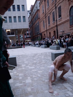 A man with knee and elbow bands is crouching on a paved urban square surrounded by modern and older brick buildings. A crowd of onlookers is gathered around the area, and another person stands in the background wearing red pants, suggesting a performance or public event.