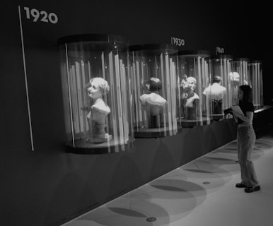 A series of mannequin busts are displayed in cylindrical glass cases, each representing different decades from the 1920s to the 1940s. A person stands observing the displays, holding a notebook and a camera, immersed in the exhibit.