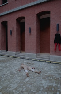 A person is lying face-up on a brick pavement in front of a red brick wall with arched doorways. The individual appears to be wearing a skin-toned outfit, and there is a black jacket with red pants hanging on the wall.