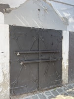 A metal door with ornate ironwork featuring two horizontal bars secured by padlocks. The structure is surrounded by a worn wall with peeling paint and some exposed concrete. A cobblestone path runs along the bottom of the image.