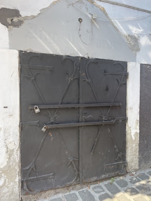 A metal door with ornate ironwork featuring two horizontal bars secured by padlocks. The structure is surrounded by a worn wall with peeling paint and some exposed concrete. A cobblestone path runs along the bottom of the image.