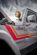 A close-up view of the side of a white Jeep Wrangler Unlimited featuring colorful decals and badges on its side. The vehicle's hood is slightly raised, revealing part of the engine area. A distinctive yellow smiley-face cover is placed over one of the vehicle's lights. Raindrops coat the surface of the vehicle, suggesting recent or ongoing rain. The sky in the background is overcast and cloudy.