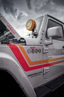 A close-up view of the side of a white Jeep Wrangler Unlimited featuring colorful decals and badges on its side. The vehicle's hood is slightly raised, revealing part of the engine area. A distinctive yellow smiley-face cover is placed over one of the vehicle's lights. Raindrops coat the surface of the vehicle, suggesting recent or ongoing rain. The sky in the background is overcast and cloudy.