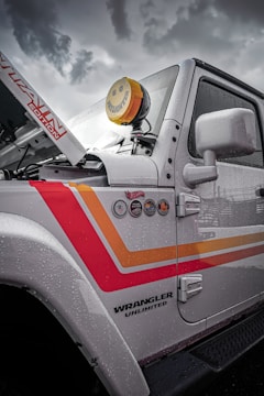 A close-up view of the side of a white Jeep Wrangler Unlimited featuring colorful decals and badges on its side. The vehicle's hood is slightly raised, revealing part of the engine area. A distinctive yellow smiley-face cover is placed over one of the vehicle's lights. Raindrops coat the surface of the vehicle, suggesting recent or ongoing rain. The sky in the background is overcast and cloudy.