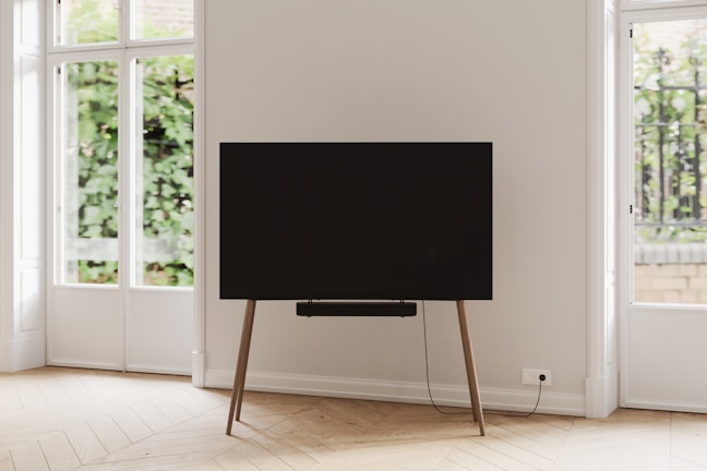 A modern and minimalistic interior space featuring a large flat-screen television mounted on a wooden stand. The room has large windows allowing natural light to illuminate the space, and outside, greenery is visible. The floor is made of light-colored wood in a herringbone pattern, and the walls are painted white.