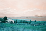 A serene countryside scene featuring a white house with a metal roof surrounded by lush greenery. The landscape extends towards a body of water, likely a lake, with mountains in the distance under a partly cloudy sky.