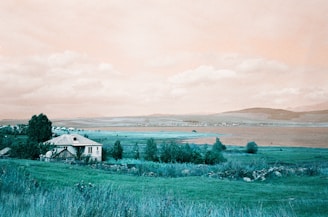A serene countryside scene featuring a white house with a metal roof surrounded by lush greenery. The landscape extends towards a body of water, likely a lake, with mountains in the distance under a partly cloudy sky.