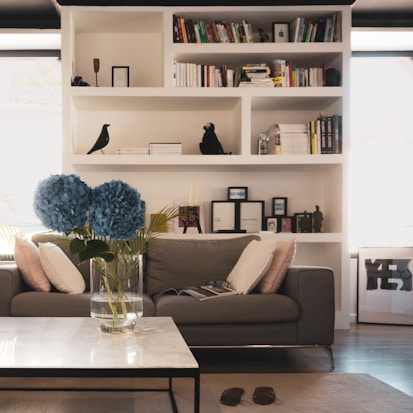 A cozy living room featuring a grey sofa with numerous cushions in front of white shelves filled with books and decorative items. A glass vase containing large blue flowers sits on a marble-topped coffee table. Sunlight is entering the room through windows on either side of the shelves. Various small objects and framed pictures are also visible on the shelves. A pair of slippers lie on the floor near the table.
