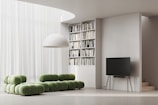 A modern living room with a minimalist design features a green modular sofa set against a backdrop of sheer white curtains. A large white dome light fixture hangs above. To the side, a tall bookshelf filled with various books is integrated into the wall. A sleek, black flat-screen TV rests on a stand with slender, wooden legs, adjacent to a small staircase.